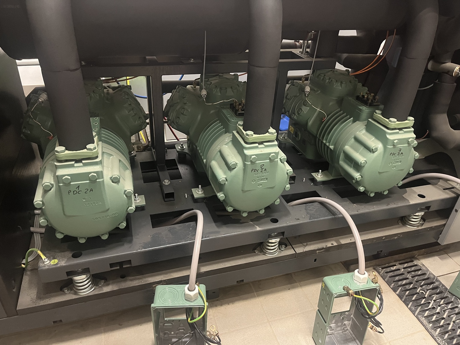 Three green industrial compressors mounted on a metal frame with connected pipes and electrical wiring in a mechanical room.