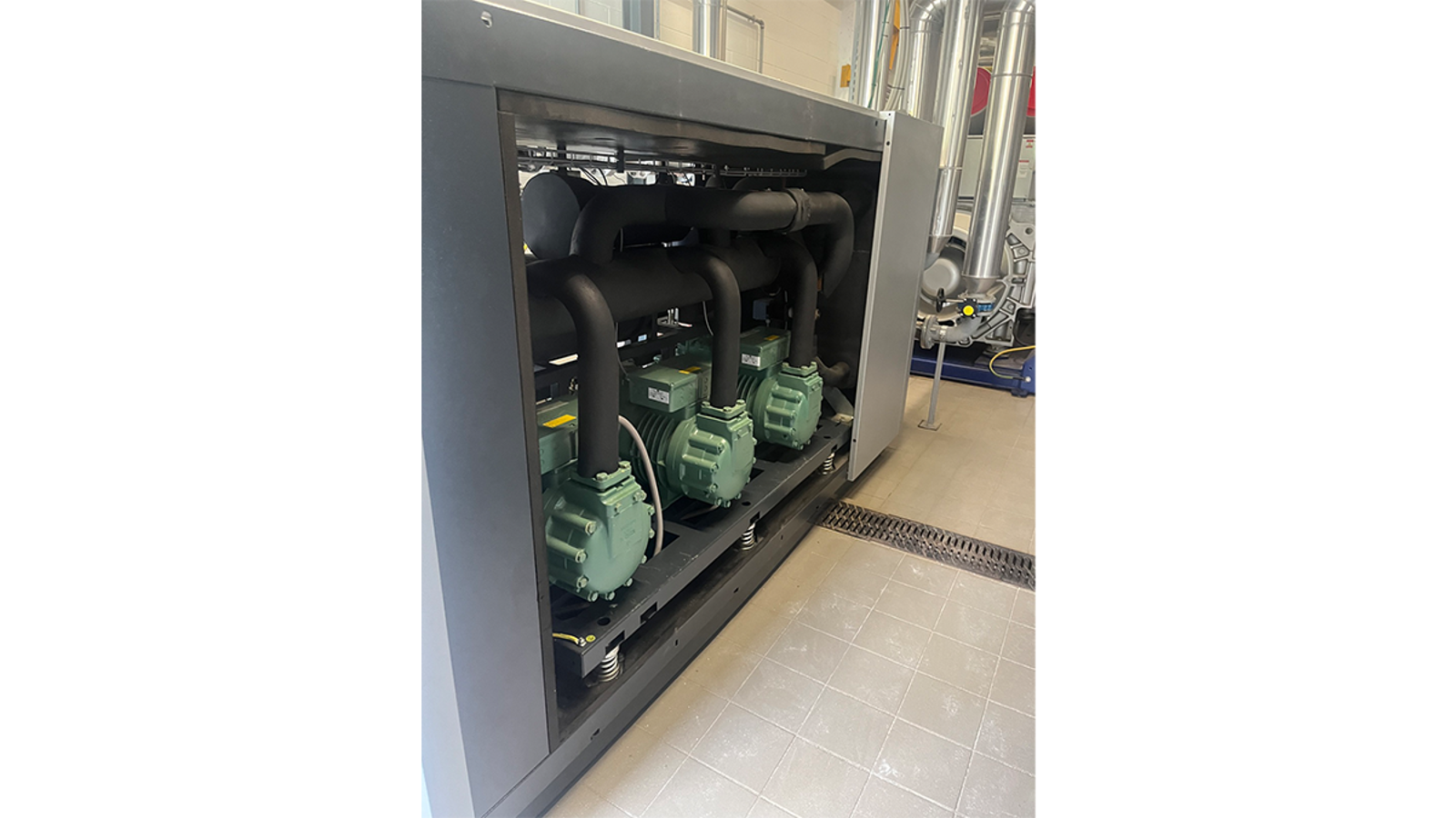 Industrial compressor unit with three green reciprocating compressors connected by black piping inside a metal enclosure, located in a facility with tiled flooring and surrounding stainless steel pipes.